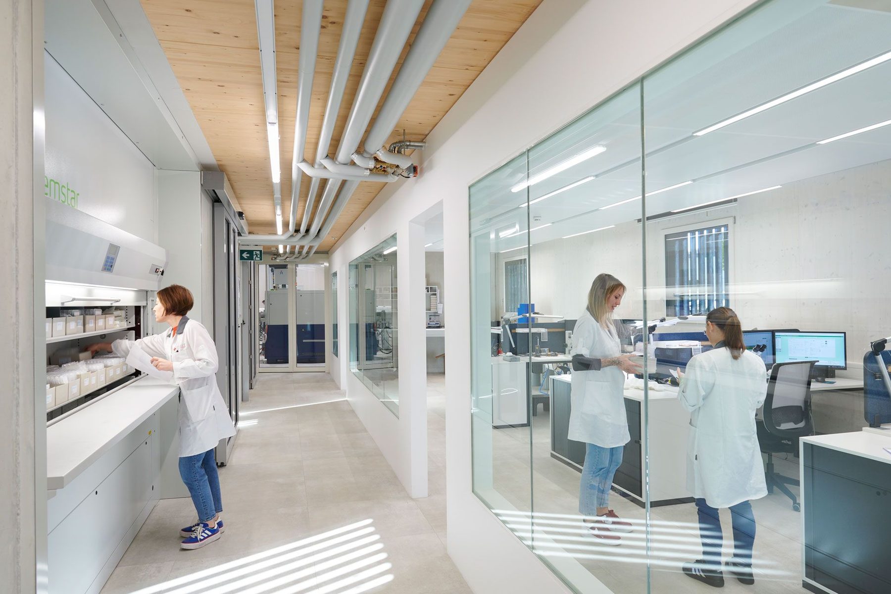 Le couloir vitré des locaux de Steulet Microtechnique, collaboratrices en blouse au laboratoire